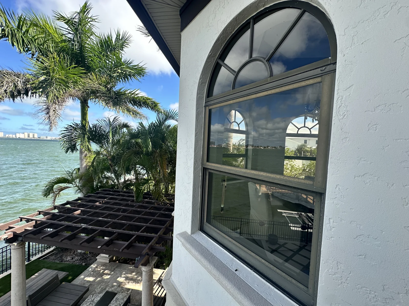 Dark-framed arched window beside a patio overlooking palm trees and the waterfront.