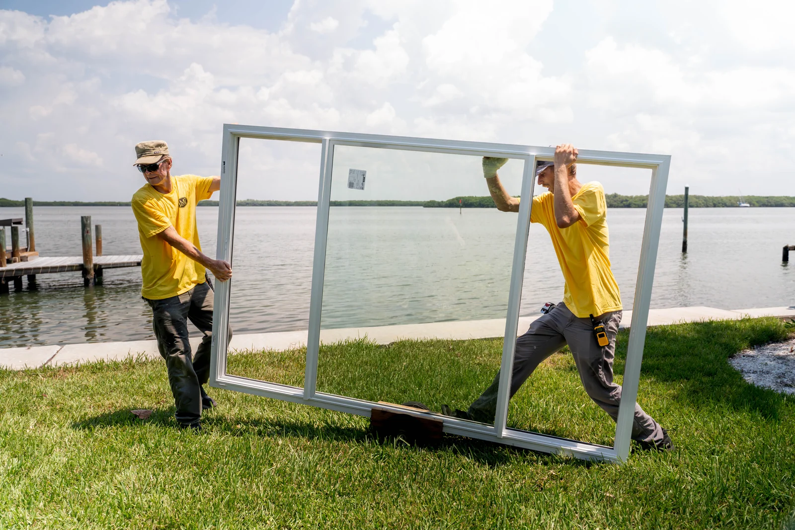 Two JMI installers carrying a large three-panel window frame beside a waterfront home.